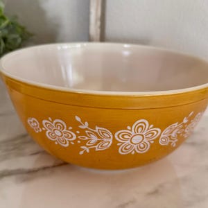 May include: A round mixing bowl with a glossy mustard yellow exterior and a smooth cream-colored interior. The outer surface features a repeating white floral pattern consisting of stylized flowers and delicate leafy vines encircling the bowl.