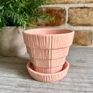 May include: A light pink ceramic plant pot with a matching saucer, featuring a textured basket-weave pattern on the exterior. The pot has a tapered cylindrical shape with a smooth rim and a consistent pastel pink glaze throughout the entire surface.