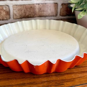 May include: A vintage pie pan with a scalloped edge. The pan has a bright orange exterior and a cream-colored interior. The pan is sitting on a wooden surface.