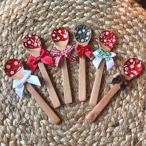 May include: Six wooden spoons with decorative red, green, and white painted designs on the spoon heads. Each spoon has a different colored bow tied around the handle, including red, white, and green.