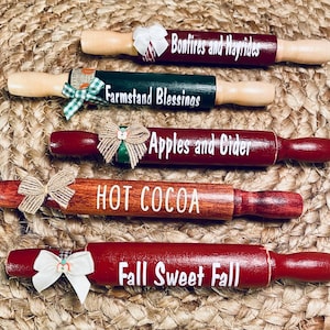 May include: Five decorative rolling pins with wooden handles and red or brown painted barrels. Each pin has white text: "Bonfires and Hayrides", "Farmstand Blessings", "Apples and Cider", "Hot Cocoa", and "Fall Sweet Fall". Each pin is decorated with a bow.