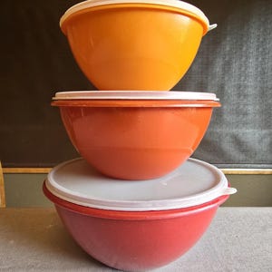 May include: Three plastic mixing bowls in shades of orange and red, stacked on top of each other. Each bowl has a white lid. The bowls are of varying sizes, with the largest at the bottom and the smallest at the top.