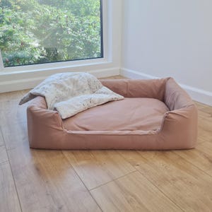 May include: A rectangular pet bed in a muted rose color, with a matching cushion and a patterned pillow. The bed is placed on a hardwood floor near a window, creating a cozy and inviting space for a pet to rest.