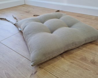 Könnte beinhalten: Ein quadratisches, hellbeige Sitzkissen mit getuftetem Design. Das Kissen hat eine strukturierte Oberfläche und liegt auf einem Holzboden. Es hat Stoffbänder zur Befestigung an einem Stuhl.