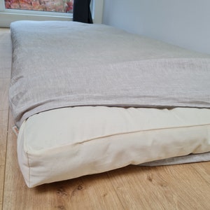 May include: A rectangular floor mattress featuring a thick, off-white cotton casing and a removable, textured light gray linen cover that partially drapes over the top. A small white fabric tag with red text is visible on the side of the mattress.