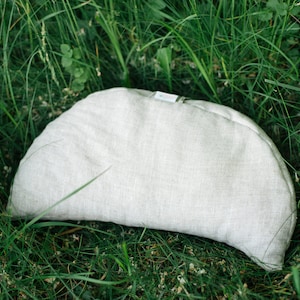 May include: A half-moon shaped pillow made of light gray linen fabric. The pillow is sitting on a bed of green grass.