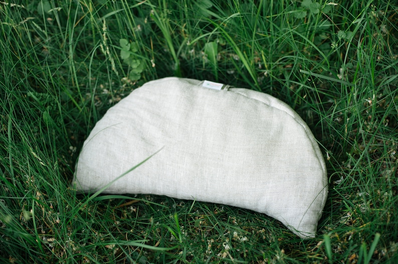 May include: A half-moon shaped pillow made of light gray linen fabric. The pillow is sitting on a bed of green grass.