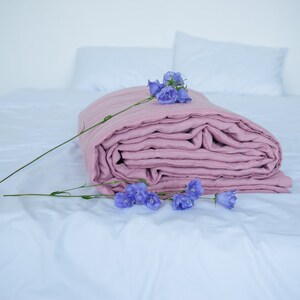 May include: A folded, dusty rose-colored linen blanket rests on a white bed, adorned with sprigs of purple flowers. The blanket is the focal point, with visible folds and a soft texture. Two white pillows are in the background.