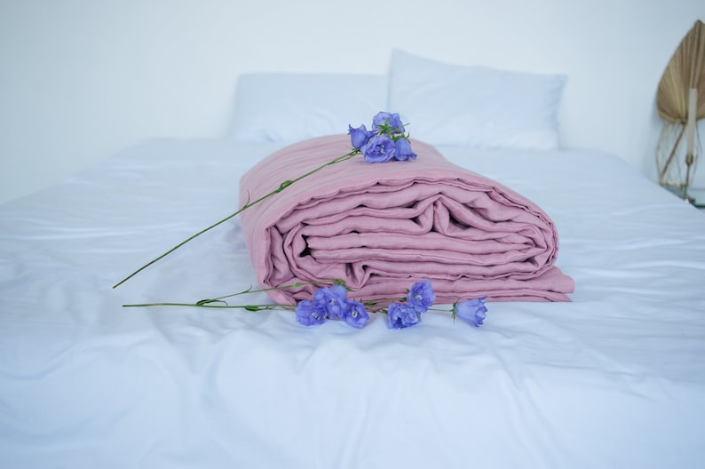 May include: A folded, dusty rose-colored linen blanket rests on a white bed, adorned with sprigs of purple flowers. The blanket is the focal point, with visible folds and a soft texture. Two white pillows are in the background.