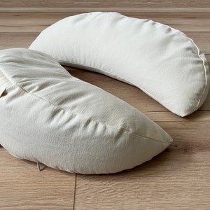 May include: Two off-white crescent-shaped pillows made of soft fabric. The pillows are placed on a light brown wooden surface.