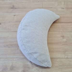 May include: A crescent-shaped pillow made of a light gray fabric with a subtle texture. The pillow is lying on a light wood floor.