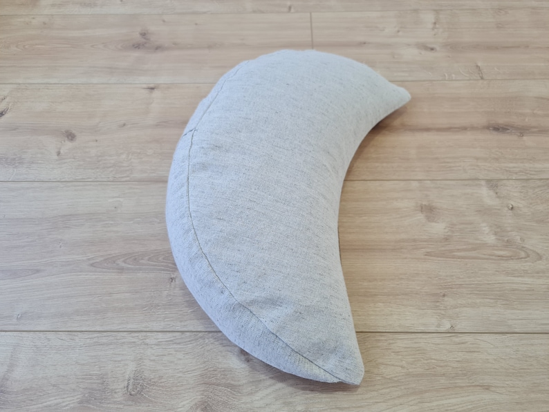 May include: A crescent-shaped pillow made of a light gray fabric with a subtle texture. The pillow is lying on a light wood floor.