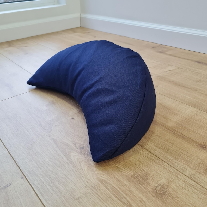 May include: A crescent moon-shaped pillow in a solid dark navy blue fabric. The pillow features a curved, semi-circular design with smooth, plain textile surfaces and stitched seams along the edges, creating a distinct, rounded, and plump form.