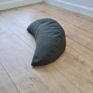 May include: A crescent-shaped meditation cushion in a solid dark olive green fabric. The pillow features a curved, half-moon design with smooth, stitched seams along the edges, providing a firm and structured shape for seated support.
