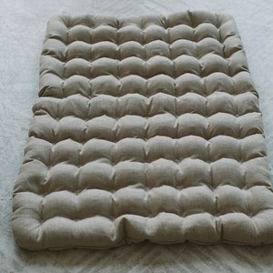 May include: A beige linen floor cushion with a tufted design. The cushion is made of a soft, natural fabric and has a comfortable, firm feel.