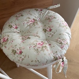 Può includere: Cuscino rotondo con un motivo floreale di rose rosa e foglie verdi su sfondo azzurro. Il cuscino è legato a una sedia bianca con lacci in tessuto coordinato. La sedia ha gambe bianche.