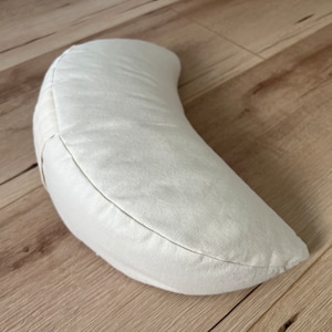 May include: A crescent-shaped meditation cushion in an off-white, cream-colored fabric. The pillow features a curved, semi-circular design with visible stitched seams along the edges, providing a structured yet soft form for seated support.