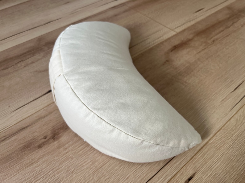 May include: A crescent-shaped meditation cushion in an off-white, cream-colored fabric. The pillow features a curved, semi-circular design with visible stitched seams along the edges, providing a structured yet soft form for seated support.