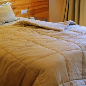 May include: A close-up of a beige quilted duvet on a bed with two pillows. The duvet is folded back to reveal the white sheets underneath. The bed is made with a wooden headboard and a bedside table with a lamp.
