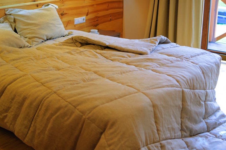 May include: A close-up of a beige quilted duvet on a bed with two pillows. The duvet is folded back to reveal the white sheets underneath. The bed is made with a wooden headboard and a bedside table with a lamp.