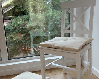 Puede incluir: Una silla de madera blanca con respaldo en forma de X y un cojín de asiento cuadrado de color blanco roto con un punto de capitoné. Un cojín de suelo cuadrado a juego, también con un punto central, se encuentra en el suelo junto a la silla.
