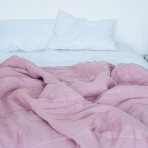 May include: A bed with a light blue fitted sheet and two white pillows. A dusty rose colored linen duvet covers the bed. A small glass side table with gold accents and decorative items sits next to the bed.
