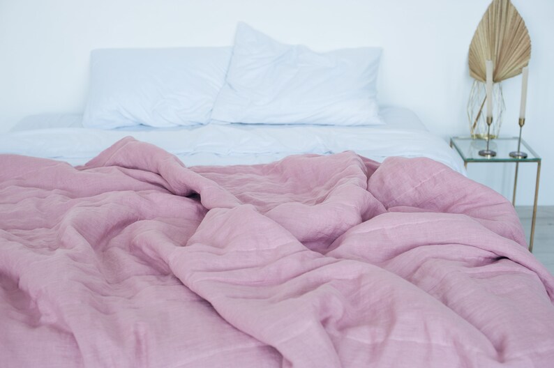 May include: A bed with a light blue fitted sheet and two white pillows. A dusty rose colored linen duvet covers the bed. A small glass side table with gold accents and decorative items sits next to the bed.