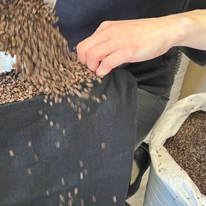 May include: A person is pouring buckwheat groats into a black fabric bag. The bag is sitting on a table and the groats are spilling out of the bag. The bag is labeled "HEMP OrganicLife".