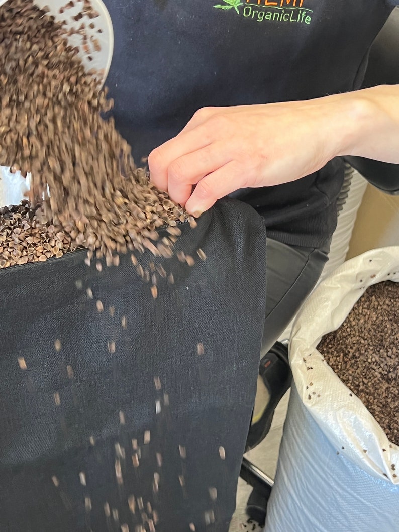 May include: A person is pouring buckwheat groats into a black fabric bag. The bag is sitting on a table and the groats are spilling out of the bag. The bag is labeled "HEMP OrganicLife".