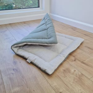 May include: A rectangular pet mat featuring a sage green top side and a light beige underside. The mat is partially folded over to reveal both colours, with a quilted texture and a small white fabric tag attached to the edge of the border.