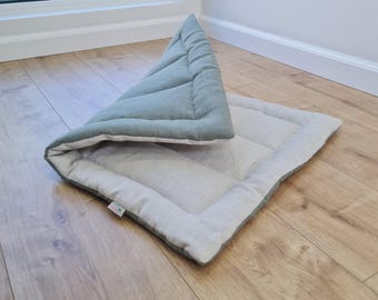 May include: A rectangular pet mat featuring a sage green top side and a light beige underside. The mat is partially folded over to reveal both colors, with a quilted texture and a small white fabric tag attached to the edge of the border.