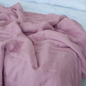 May include: A dusty rose-colored linen duvet cover draped over a bed. The cover has a textured appearance and is partially covering a white bed sheet. The bedding is arranged on a bed with a white headboard and light-colored flooring.
