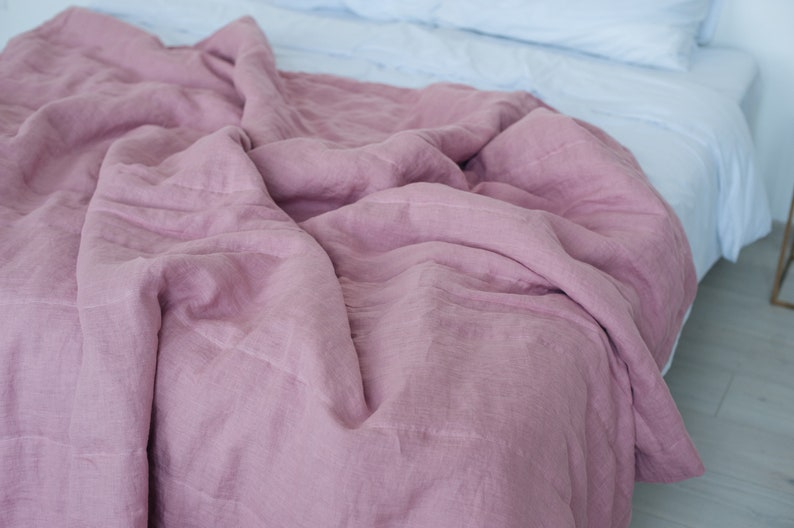 May include: A dusty rose-colored linen duvet cover draped over a bed. The cover has a textured appearance and is partially covering a white bed sheet. The bedding is arranged on a bed with a white headboard and light-colored flooring.