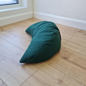 May include: A crescent-shaped meditation cushion in a solid dark forest green fabric. The pillow features a curved, ergonomic design with visible seams along the edges, providing a structured form that rests on its side on a flat surface.
