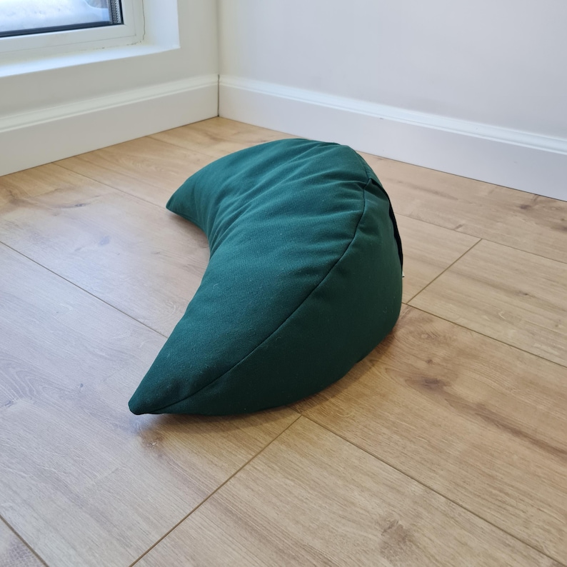 May include: A crescent-shaped meditation cushion in a solid dark forest green fabric. The pillow features a curved, ergonomic design with visible seams along the edges, providing a structured form that rests on its side on a flat surface.