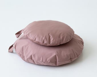 May include: Two round, dusty rose-colored cushions are stacked on top of each other. Each cushion has a fabric loop attached. The cushions are made of a soft, smooth material and are set against a white background.