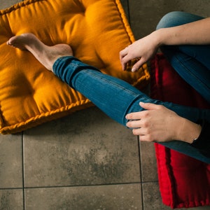 Puede incluir: Una persona sentada en un cojín de suelo rojo y amarillo con las piernas cruzadas. Lleva pantalones vaqueros azules y una camisa negra de manga larga.