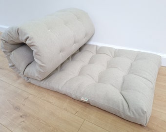 Puede incluir: Un colchón futón beige con parte superior acolchada y diseño capitoné. El colchón está enrollado y apoyado en un suelo de madera.