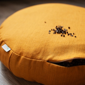 May include: A round mustard yellow meditation cushion with a zipper opening on the side. The cushion is filled with buckwheat hulls, which are visible through the opening.