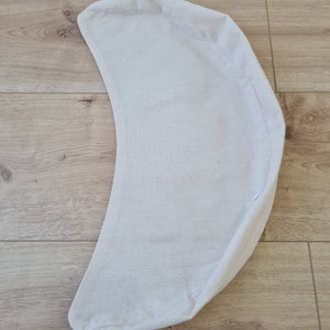May include: A white crescent-shaped cushion made of fabric. The cushion is lying flat on a wooden surface.