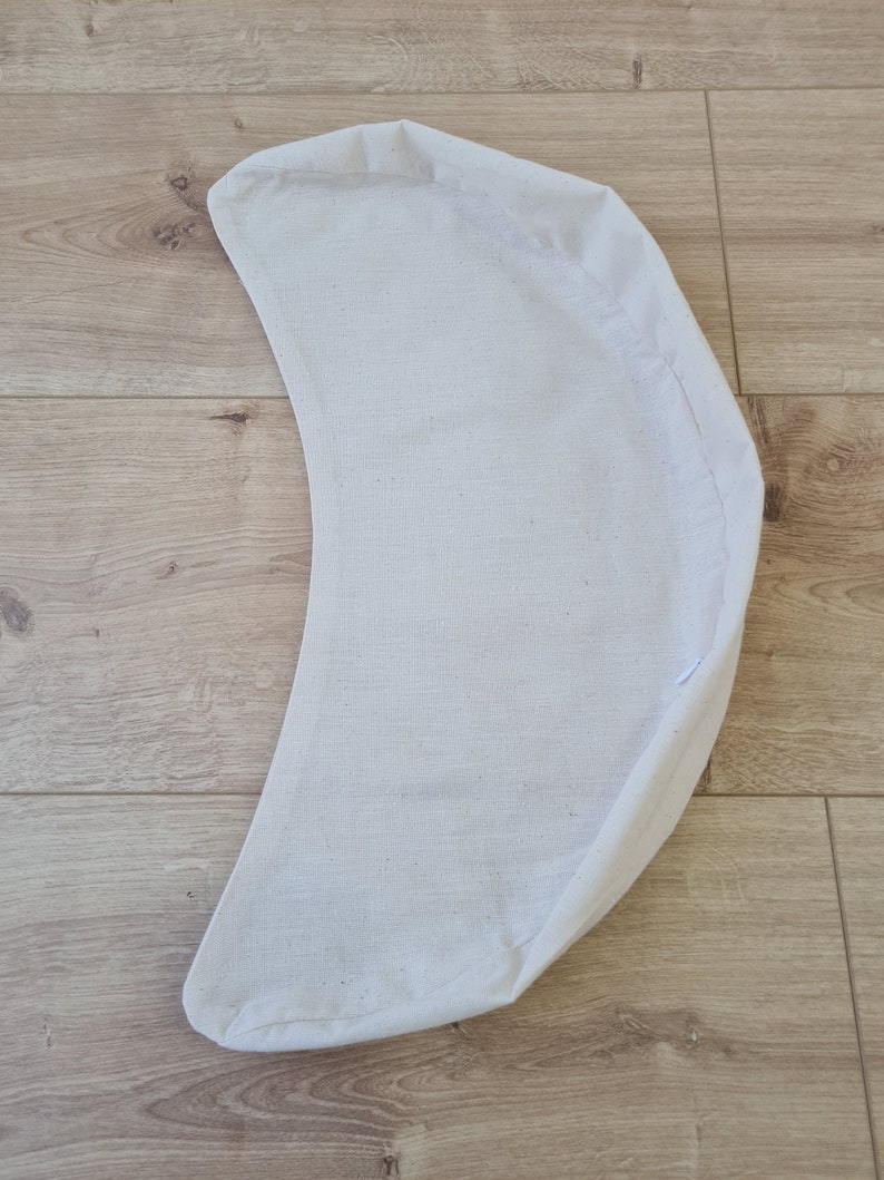 May include: A white crescent-shaped cushion made of fabric. The cushion is lying flat on a wooden surface.