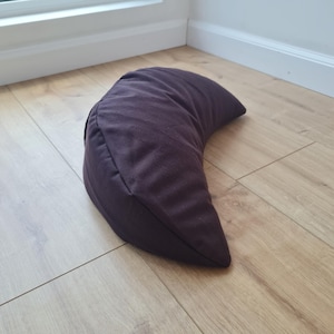 May include: A crescent-shaped meditation cushion in a solid dark brown fabric. The pillow features a curved, ergonomic design with visible seams along the edges, providing a structured form that tapers to points at both ends of the moon shape.