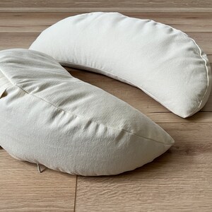 May include: Two off-white crescent-shaped pillows made of a soft, textured fabric. The pillows are placed on a light brown wooden surface.