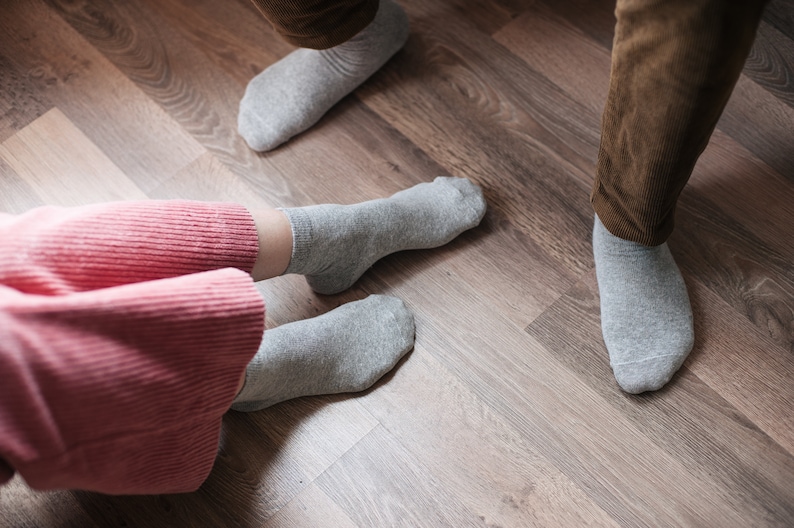 May include: Two pairs of grey socks on a wooden floor. One pair is on a person's feet, the other pair is on the floor.