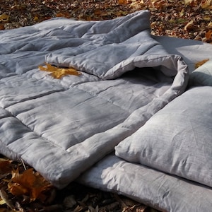 May include: A grey linen duvet cover with a matching pillow and a quilted pillow. The bedding is laid out on a bed of fallen leaves.