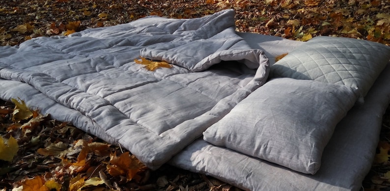 May include: A grey linen duvet cover with a matching pillow and a quilted pillow. The bedding is laid out on a bed of fallen leaves.