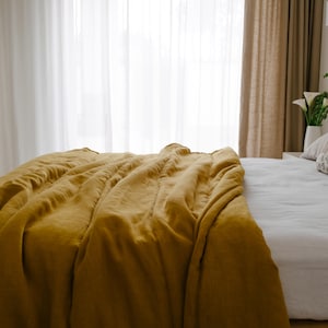 Könnte beinhalten: Ein Bett mit einer senfgelben Decke. Das Bett hat ein weißes Laken und ein gemustertes Kissen. Transparente weiße Vorhänge und ein gewebtes Kopfteil sind im Hintergrund zu sehen.