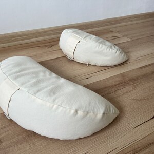May include: Two off-white crescent-shaped meditation cushions with a white strap on each.