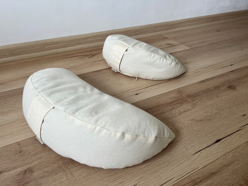 May include: Two off-white crescent-shaped meditation cushions with a white strap on each.