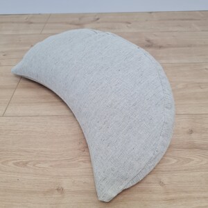 May include: A crescent-shaped pillow made of a light gray linen fabric. The pillow is lying on a light brown wooden floor.